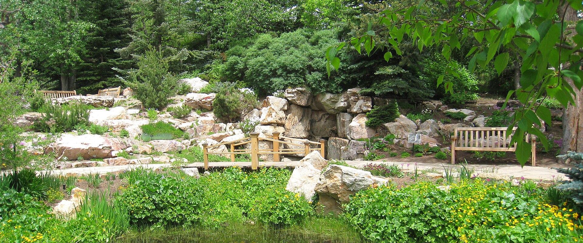 A landscaped garden scene featuring a small pond in the foreground, a wooden footbridge crossing a stream, and a rocky hillside with benches and various trees and shrubs.