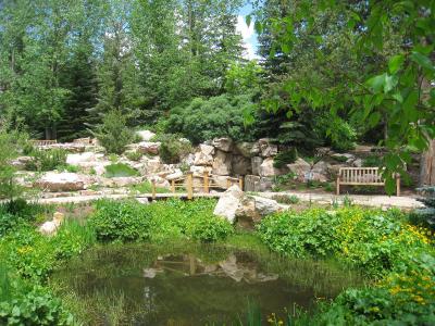 A landscaped garden scene featuring a small pond in the foreground, a wooden footbridge crossing a stream, and a rocky hillside with benches and various trees and shrubs.