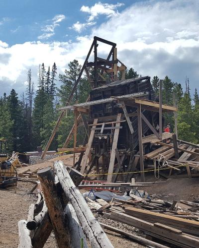 A large wooden mining headframe structure under active restoration or construction, with a yellow front-end loader, piles of lumber, and workers visible in a clearing surrounded by pine trees.