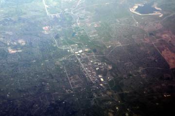 An aerial photograph showing a sprawling suburban landscape with residential developments, roads, and patches of greenery, likely taken from an airplane.