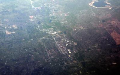 An aerial photograph showing a sprawling suburban landscape with residential developments, roads, and patches of greenery, likely taken from an airplane.