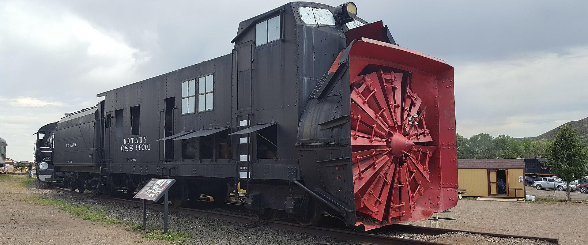 A large black rotary snowplow locomotive with a prominent red cutting wheel on the front, sitting on tracks outdoors with an informational plaque nearby.