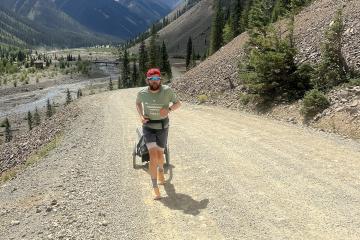 A man wearing a red cap and sunglasses runs on a gravel road pulling a small cart, with mountains and a river in the background.