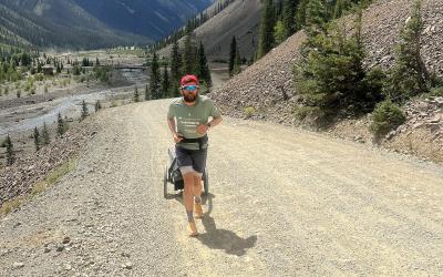 A man wearing a red cap and sunglasses runs on a gravel road pulling a small cart, with mountains and a river in the background.