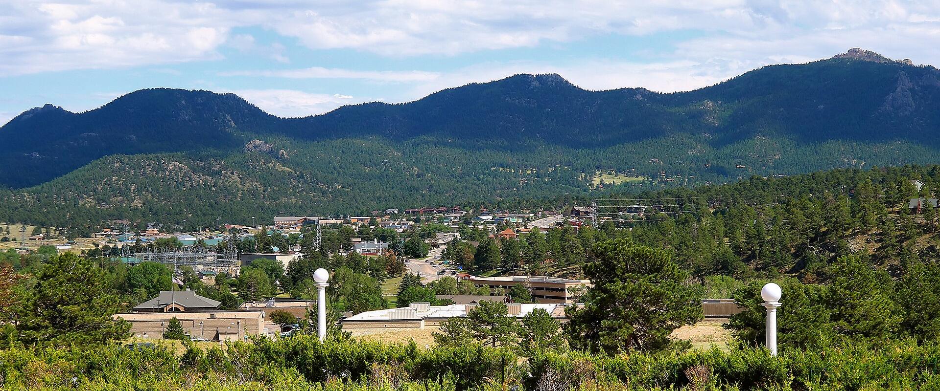 A scenic view of a town nestled in a valley surrounded by forested mountains, featuring buildings, roads, and two white spherical lamp posts in the foreground.