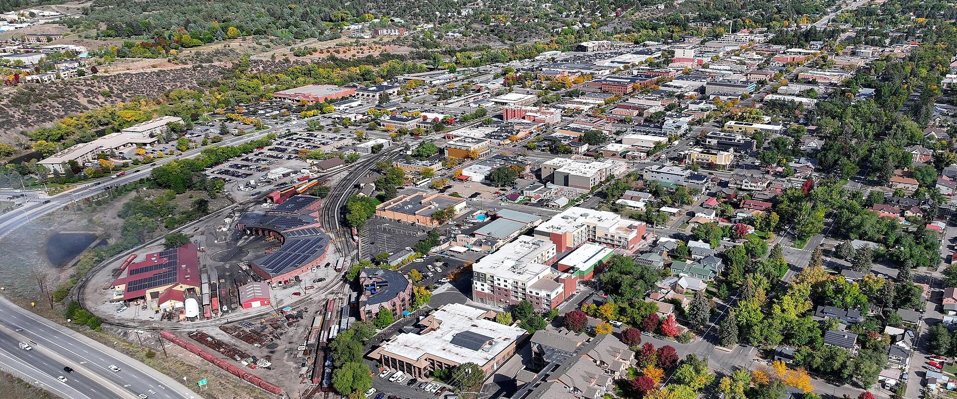 An aerial view of a town nestled in a valley with mountains in the background, featuring a prominent red brick roundhouse and rail yard in the lower left foreground.