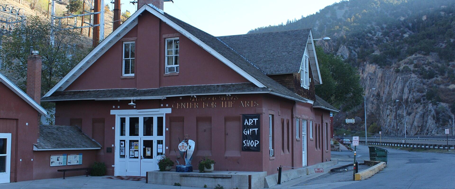 A reddish-brown building with white trim, identified by gold lettering on the facade as the "Glenwood Springs Center for the Arts" and a chalkboard sign advertising an "ART GIFT SHOP," situated next to a road with hills in the background.