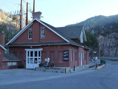 A reddish-brown building with white trim, identified by gold lettering on the facade as the "Glenwood Springs Center for the Arts" and a chalkboard sign advertising an "ART GIFT SHOP," situated next to a road with hills in the background.