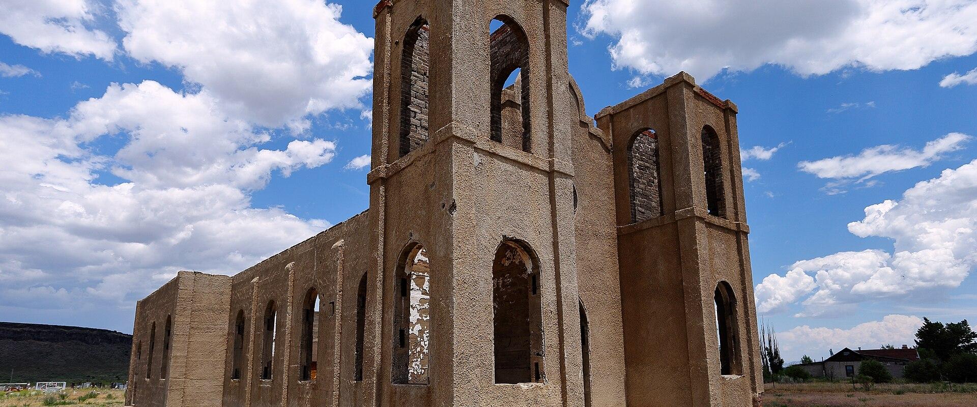 A ruined adobe church with two bell towers and arched openings stands in a grassy field under a cloudy blue sky.
