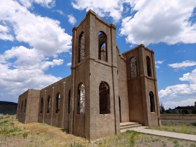 A ruined adobe church with two bell towers and arched openings stands in a grassy field under a cloudy blue sky.
