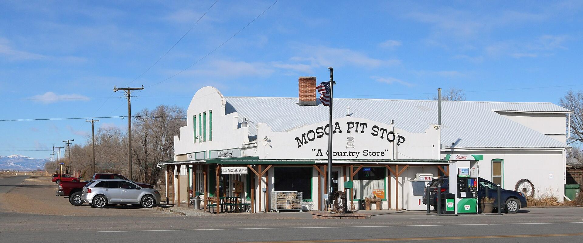 A white, single-story commercial building with a metal roof, identified by large painted lettering on the facade as "MOSCA PIT STOP 'A Country Store'", situated next to a paved road with vehicles and mountains in the background.