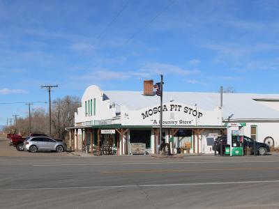 A white, single-story commercial building with a metal roof, identified by large painted lettering on the facade as "MOSCA PIT STOP 'A Country Store'", situated next to a paved road with vehicles and mountains in the background.