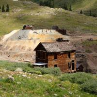 A weathered wooden building stands in the foreground of a high-altitude mountain landscape, with a large, excavated tailings pile and additional ruins visible on the hillside behind it.