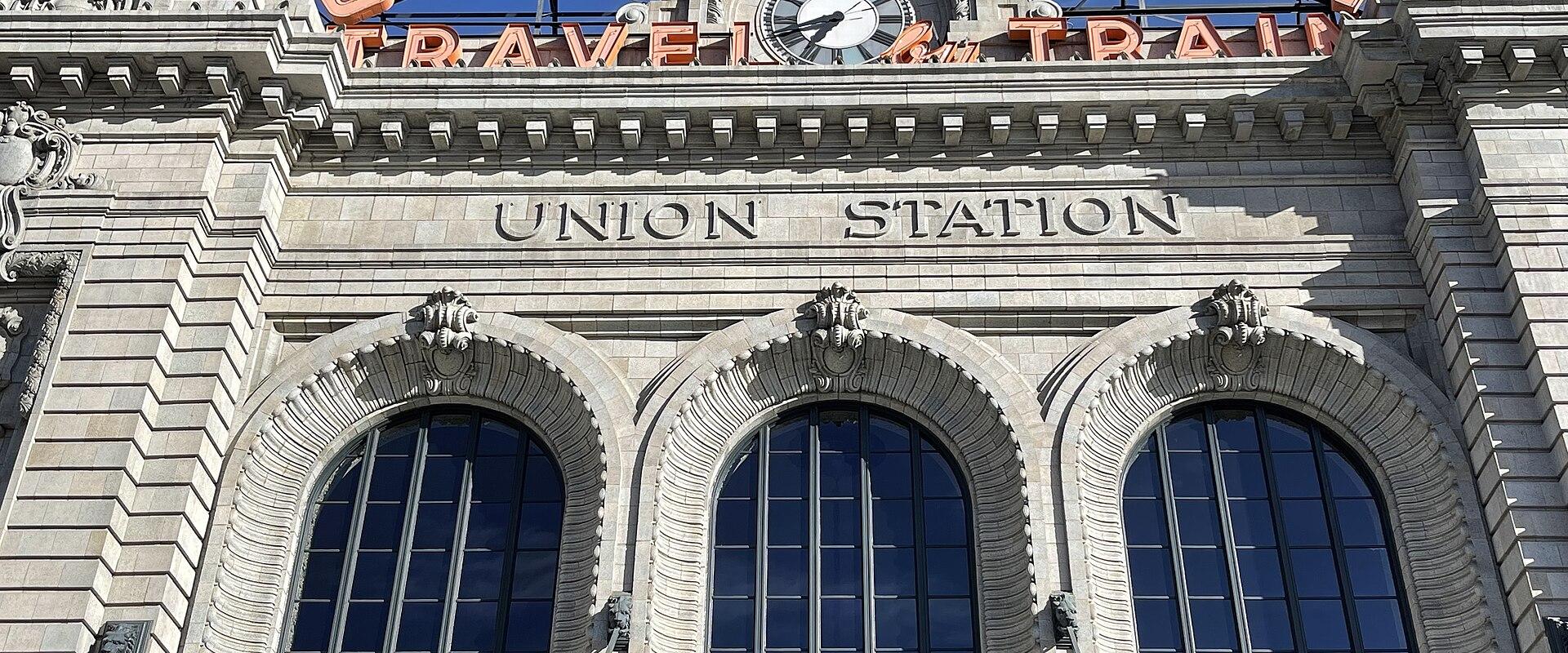 A low-angle shot of the Beaux-Arts facade of a large stone building featuring three arched windows, a central clock, and prominent signage reading "UNION STATION" in both carved stone and large orange neon letters.