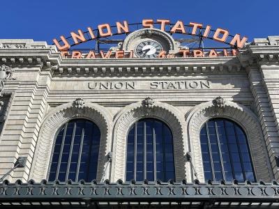 A low-angle shot of the Beaux-Arts facade of a large stone building featuring three arched windows, a central clock, and prominent signage reading "UNION STATION" in both carved stone and large orange neon letters.