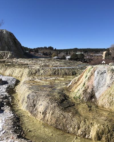 The image shows a series of terraced, mineral-rich hot spring pools with travertine formations, set against a backdrop of a small town and forested hills under a clear blue sky.