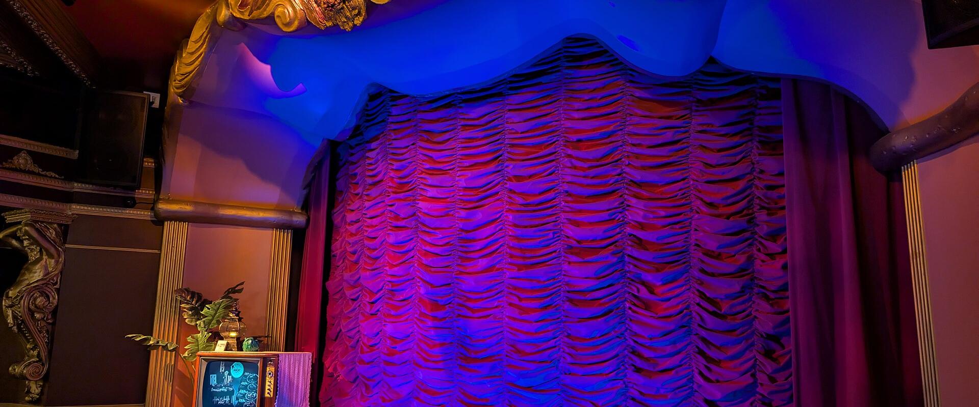 An indoor stage featuring a ruffled curtain illuminated by blue and purple lighting, with an ornate gold proscenium arch and a vintage television set on the stage floor.
