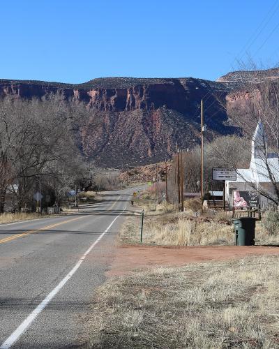 A paved two-lane road leading towards a large mesa in the distance, with a green road sign on the right reading "Gateway" and a white church visible nearby.