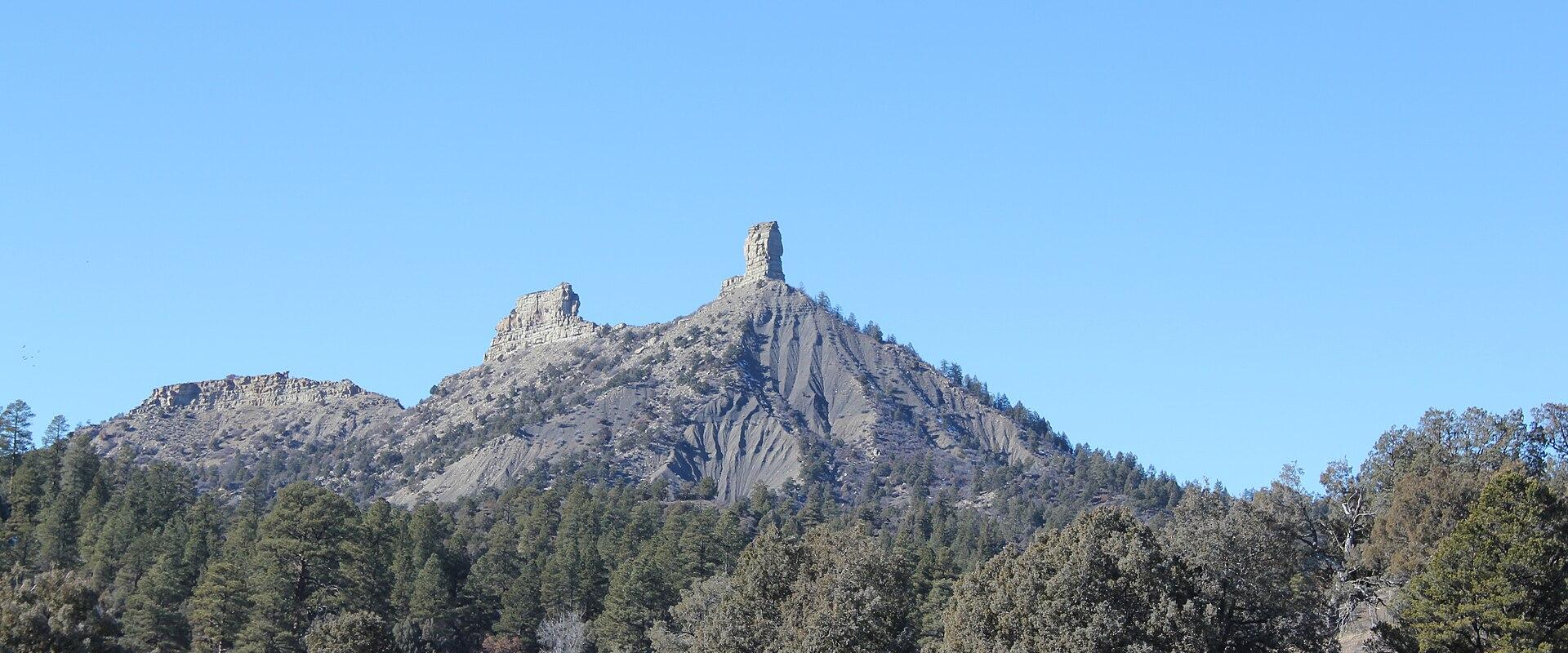 A landscape photograph showing a prominent, tall, isolated rock spire (chimney) atop a mesa, surrounded by a forest of coniferous trees under a clear blue sky.