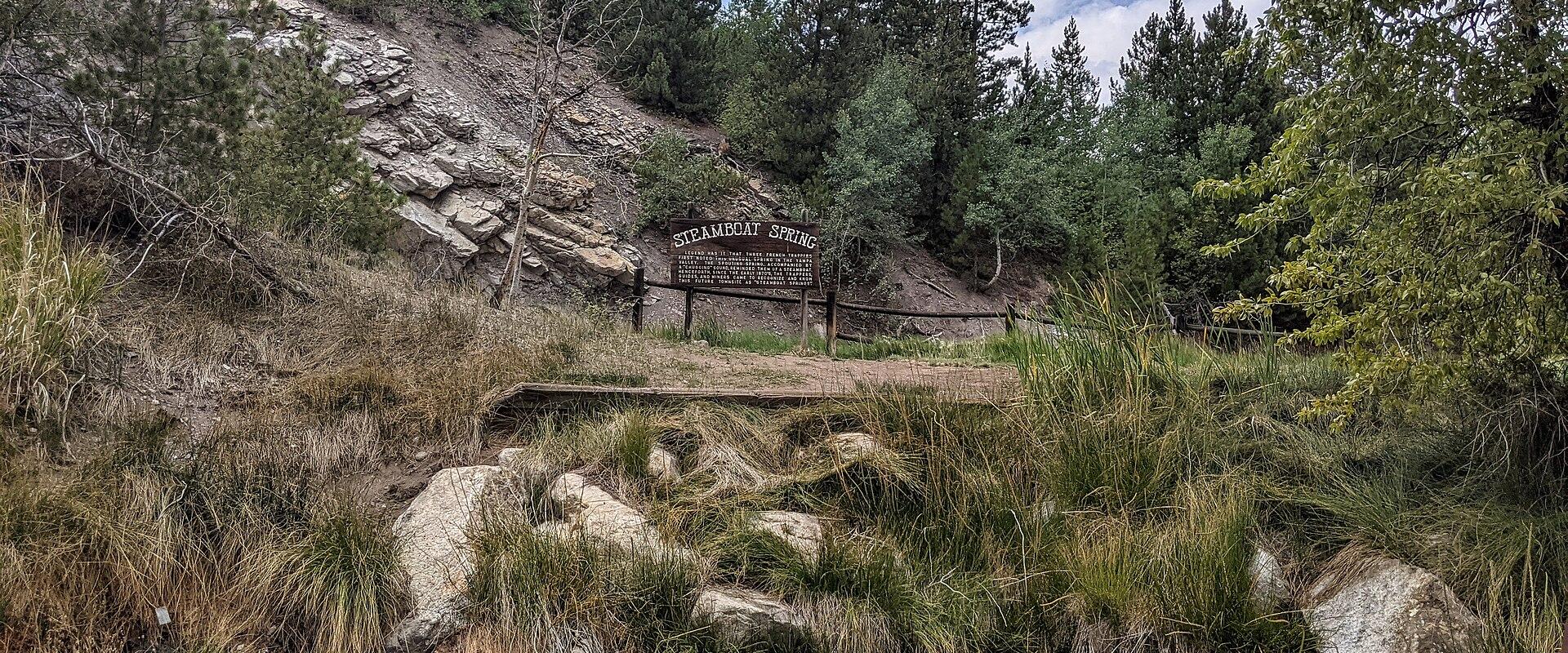A wooden boardwalk leads to a bubbling hot spring pool in a grassy, rocky area with a wooden sign reading "STEAMBOAT SPRING" in the background.