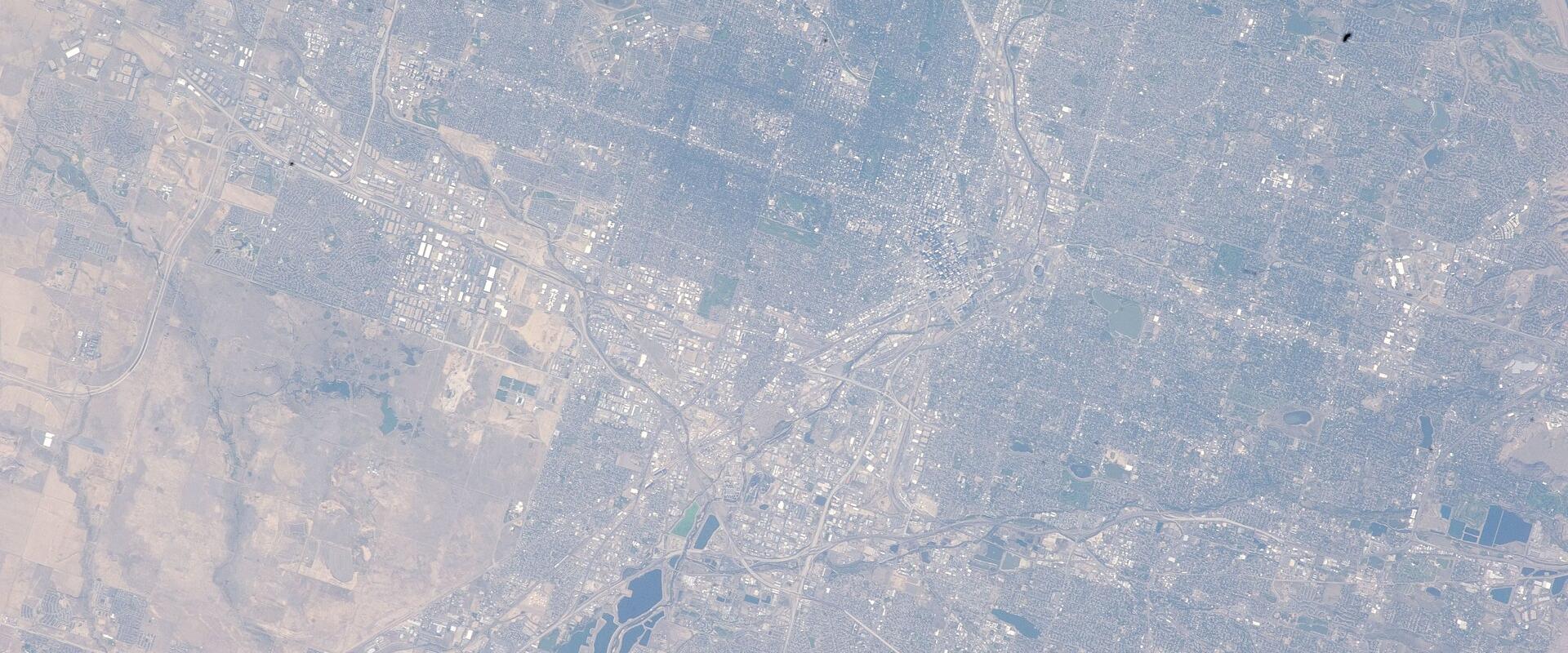 A satellite/aerial view of a sprawling urban area with a grid-like street pattern, agricultural fields, and several dark blue rectangular reservoirs or ponds in the lower-left quadrant.