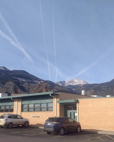 A single-story brick school building with a parking lot in the foreground and a large mountain range in the background.