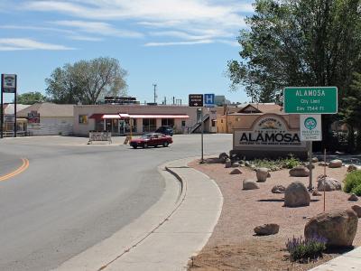 A street corner view of the entrance to the city of Alamosa, Colorado, featuring a large "Welcome to the City of ALAMOSA" sign, a green city limit sign, and commercial buildings in the background.