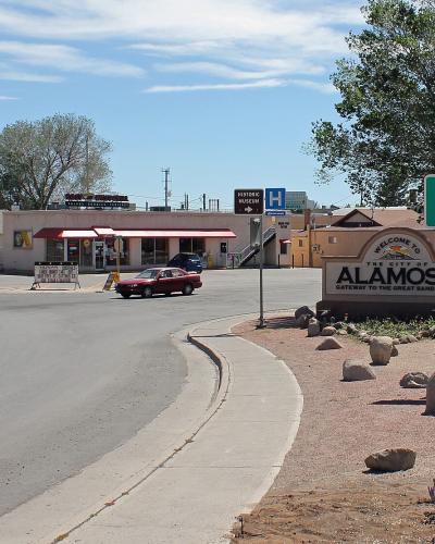 A street corner view of the entrance to the city of Alamosa, Colorado, featuring a large "Welcome to the City of ALAMOSA" sign, a green city limit sign, and commercial buildings in the background.
