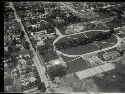 A black-and-white aerial photograph showing a large institutional campus with a central oval green space, surrounding buildings, and adjacent residential streets.
