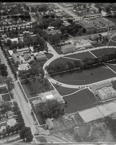 A black-and-white aerial photograph showing a large institutional campus with a central oval green space, surrounding buildings, and adjacent residential streets.