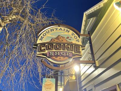 A close-up of an illuminated hanging sign for the "Mountain Top Cookie Shop" attached to the exterior of a building at night, with bare tree branches visible against a dark blue sky.