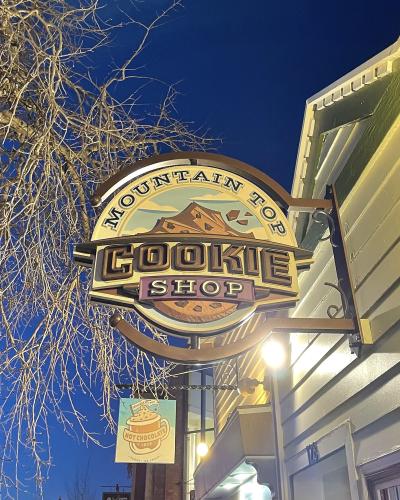 A close-up of an illuminated hanging sign for the "Mountain Top Cookie Shop" attached to the exterior of a building at night, with bare tree branches visible against a dark blue sky.