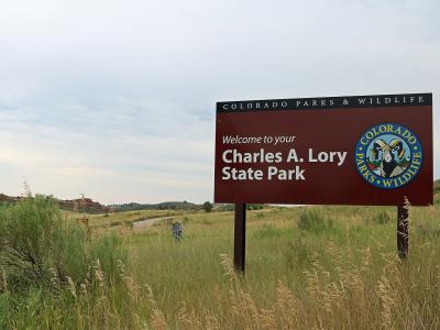 A large brown welcome sign for "Charles A. Lory State Park" under the "Colorado Parks & Wildlife" banner, set against a backdrop of grassy hills and a dirt road.