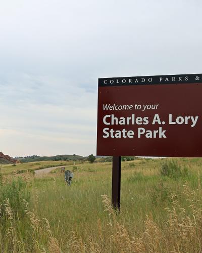 A large brown welcome sign for "Charles A. Lory State Park" under the "Colorado Parks & Wildlife" banner, set against a backdrop of grassy hills and a dirt road.