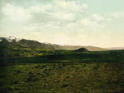 A landscape photograph showing a rolling, grassy foreground leading to a range of mountains in the distance, with a small, dark, isolated hill or butte visible in the middle ground.
