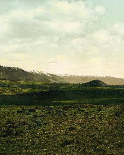A landscape photograph showing a rolling, grassy foreground leading to a range of mountains in the distance, with a small, dark, isolated hill or butte visible in the middle ground.