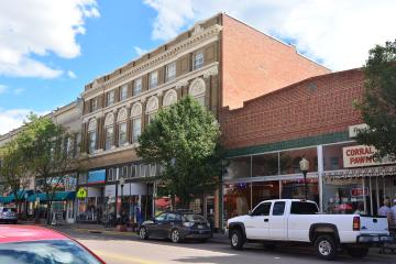 A street-level view of a historic downtown district featuring a prominent multi-story brick building with ornate white stone detailing and arched windows, alongside storefronts including one with a "CORRAL PAWN" sign.