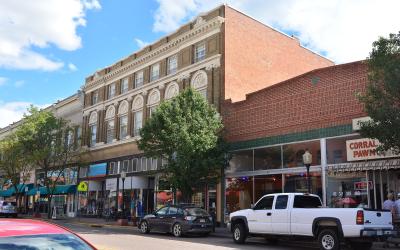 A street-level view of a historic downtown district featuring a prominent multi-story brick building with ornate white stone detailing and arched windows, alongside storefronts including one with a "CORRAL PAWN" sign.