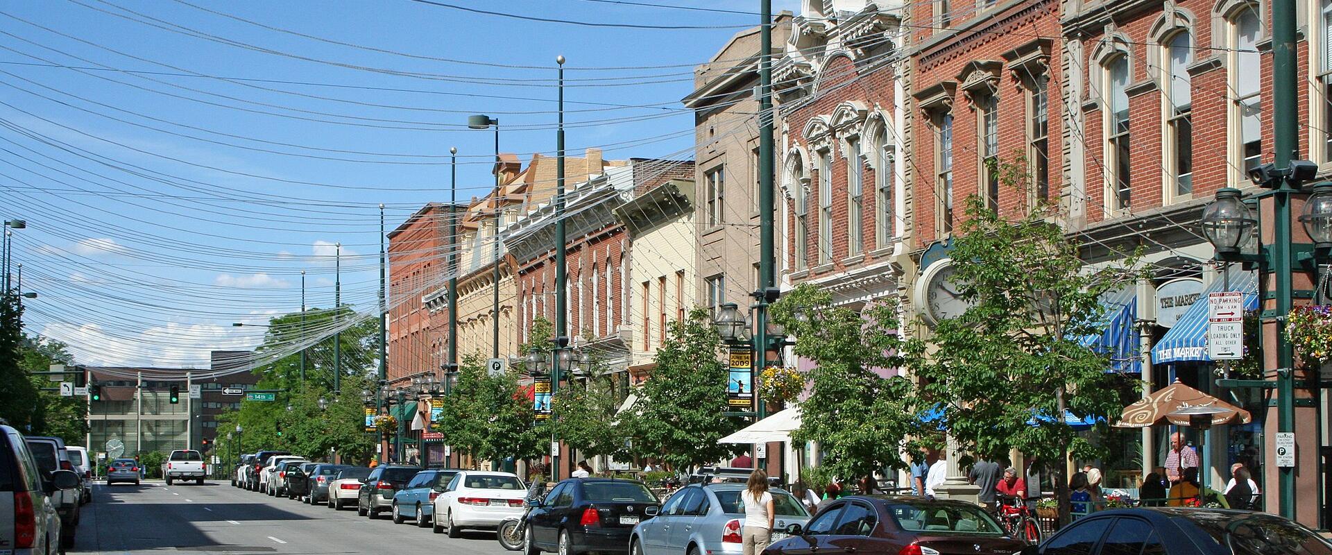 A street-level view of a historic downtown district featuring rows of parked cars along a paved road, flanked by multi-story 19th-century brick buildings with storefronts, awnings, and street lamps under a blue sky.