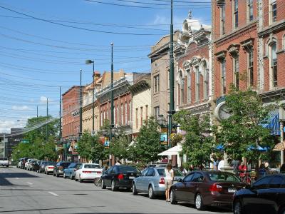 A street-level view of a historic downtown district featuring rows of parked cars along a paved road, flanked by multi-story 19th-century brick buildings with storefronts, awnings, and street lamps under a blue sky.