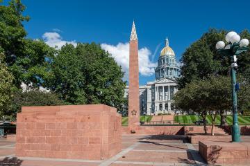 The image shows the Colorado State Capitol building with its distinctive gold dome in the background, a tall reddish-brown obelisk (the Colorado Fallen Heroes Memorial), and a matching stone memorial wall in the foreground inscribed with "COLORADO FA