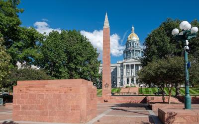 The image shows the Colorado State Capitol building with its distinctive gold dome in the background, a tall reddish-brown obelisk (the Colorado Fallen Heroes Memorial), and a matching stone memorial wall in the foreground inscribed with "COLORADO FA
