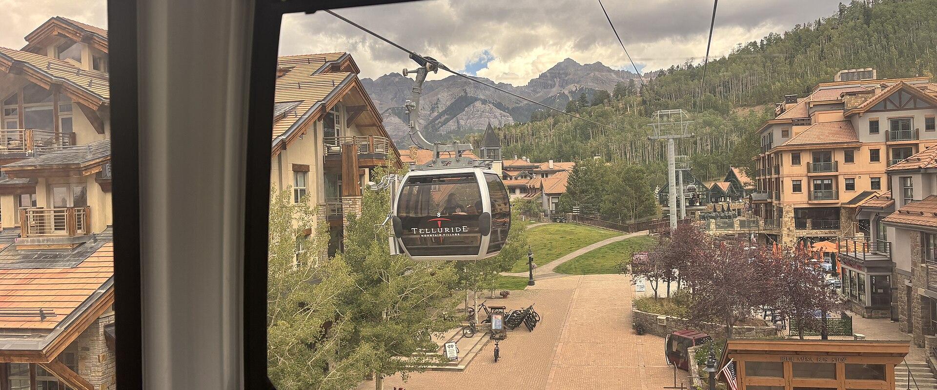 A view from inside a gondola looking out at another gondola car labeled "TELLURIDE" suspended over a paved plaza with alpine-style buildings and mountains in the background.
