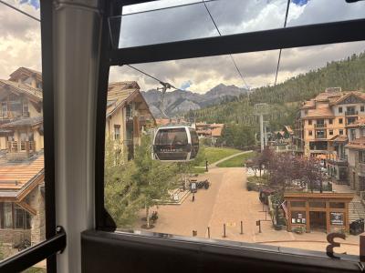 A view from inside a gondola looking out at another gondola car labeled "TELLURIDE" suspended over a paved plaza with alpine-style buildings and mountains in the background.