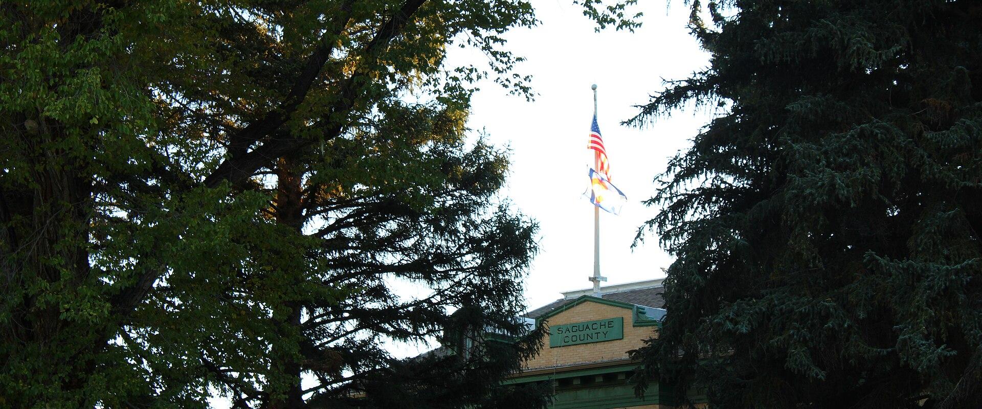 A view of the upper portion of a historic building framed by trees, featuring a flagpole with American and Colorado flags and a green sign reading "SAGUACHE COUNTY".
