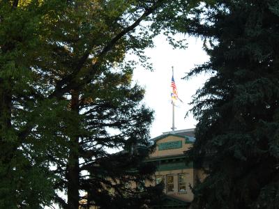 A view of the upper portion of a historic building framed by trees, featuring a flagpole with American and Colorado flags and a green sign reading "SAGUACHE COUNTY".
