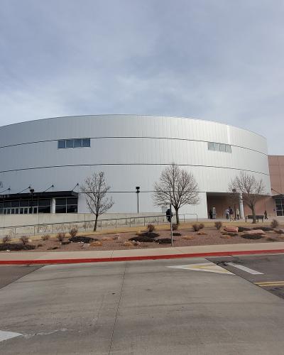 A large, modern, white cylindrical arena building with a logo and the text "WORLD ARENA" visible on its facade, situated next to a parking lot and sidewalk under a cloudy sky.