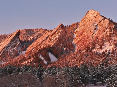 A landscape photograph showing the distinctive tilted slabs of sandstone known as the Flatirons, illuminated by warm sunlight with snow covering the slopes and trees.