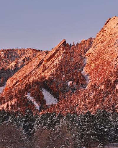 A landscape photograph showing the distinctive tilted slabs of sandstone known as the Flatirons, illuminated by warm sunlight with snow covering the slopes and trees.