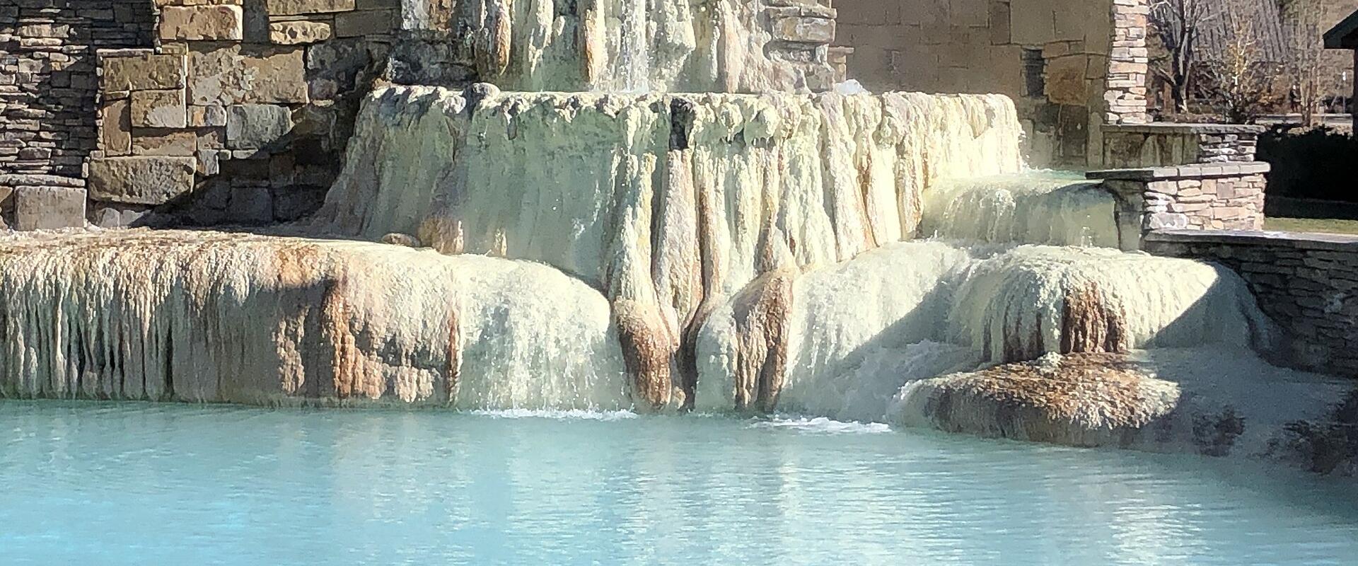 A tiered, man-made travertine waterfall structure with mineral deposits cascading into a turquoise pool, surrounded by stone retaining walls.